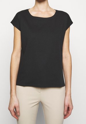 Woman wearing a plain black short-sleeve blouse paired with light beige fitted pants, standing against a neutral background.