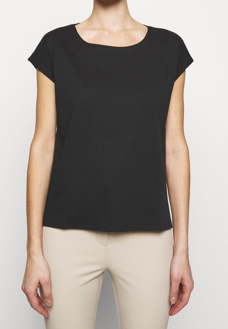 Vrouw draagt een effen zwarte kortärmelig blouse, gecombineerd met licht beige aangesloten broek, staand tegen een neutrale achtergrond.