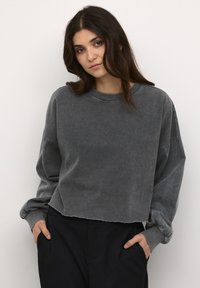 Pilki trumpi megztinis, pagamintas iš minkštos, tekstūruotos medžiagos. Yra nukritę pečių siūlai, raukšlėtos rankogaliai ir neapdirbtas kraštas. Suderintas su juodomis kelnėmis.