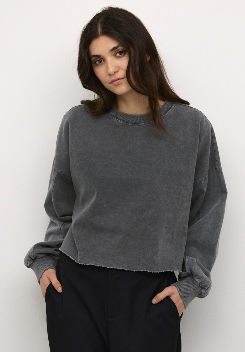 Pilki trumpi megztinis, pagamintas iš minkštos, tekstūruotos medžiagos. Yra nukritę pečių siūlai, raukšlėtos rankogaliai ir neapdirbtas kraštas. Suderintas su juodomis kelnėmis.