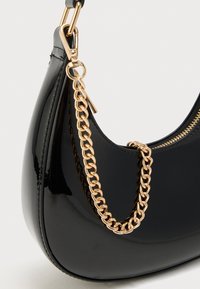 Sac à main noir brillant avec une bandoulière chaîne dorée et un détail de fermeture éclair visible sur fond blanc.