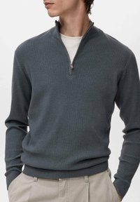 Dunkelgrauer, gerippter Pullover mit Reißverschlusskragen, aus strukturiertem Stoff. Lange Ärmel und taillierter Saum. Über einem weißen Hemd getragen.