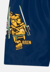 Marineblaue Shorts mit einem grafischen Motiv eines gelben Ninja-Charakters, der grüne Schwerter wieldet, mit detaillierten Designelementen und einem Namensschild "ARIN."