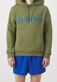 Olivově zelená mikina s kapucí a modrou vyšitou značkou "Études", přední kapsa, kapuce se stahovací šňůrkou, žebrované manžety, v kombinaci s modrými šortkami.