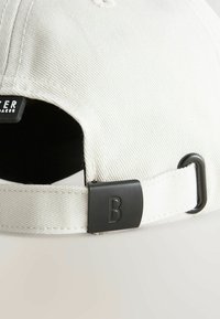 Casquette de baseball en tissu blanc avec sangle ajustable et boucle noire ornée d'une lettre "B" embossée sur un fond clair.
