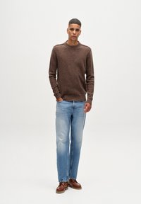 Brauner Strickpullover mit rundem Ausschnitt, kombiniert mit hellblauen Gerade geschnittenen Jeans und braunen Schuhen, vor einem schlichten weißen Hintergrund stehend.