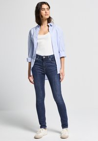 Camisa de botón azul claro sobre una camiseta blanca sin mangas, combinada con jeans ajustados azul oscuro y zapatillas beige. Tejidos suaves, corte estándar.