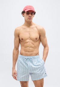 Άνδρας χωρίς πουκάμισο που φοράει κόκκινο καπέλο και ανοιχτό μπλε ριγέ μαγιό, με τα χέρια στις τσέπες, στέκεται μπροστά σε απλό λευκό φόντο.