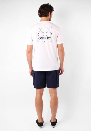 T-shirt en coton blanc avec un imprimé graphique bleu portant "OXBOW" et des éléments décoratifs au dos ; associé à un short cargo bleu marine.