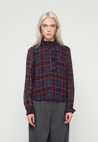 Fiatal nő, hosszú szőke hajjal, bordó és kék kockás, hosszú ujjú blúzban és szürke, széles szárú nadrágban áll egyenesen.