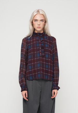 Jovem mulher com cabelo louro longo, vestindo uma blusa de manga longa xadrez em burgundy e azul e calças largas cinzas, em pé.