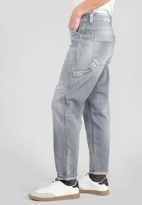 Persona vestida con pantalones de trabajo de mezclilla gris claro con múltiples bolsillos y zapatillas blancas con suelas de goma, de pie de lado.