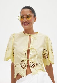 Rumena bluzica z vezenimi čipkastimi podrobnostmi, kratkimi nabranimi rokavi in sprednjim vezanjem. Ima valovit rob in zapletene vzorce.