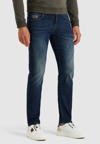 Tummansiniset farkut slim fit -leikkauksella, joissa on vaaleaa kulumista, klassinen viiden taskun muotoilu ja ruskeat ompeluyksityiskohdat. Vaatteet on yhdistetty valkoisiin tennareihin.
