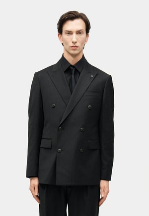 Homme portant une veste noire croisée, une chemise noire assortie et une cravate noire, se tenant devant un fond clair uni.