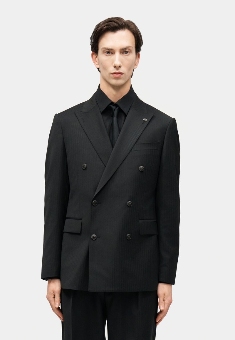 Άνδρας φοράει διπλόπετο μαύρο σακάκι με αντίστοιχο μαύρο πουκάμισο και γραβάτα, στέκεται μπροστά σε απλό φωτεινό φόντο.