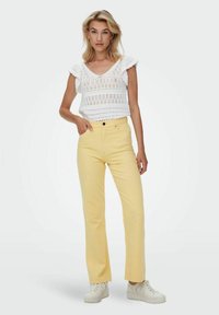Blusa blanca de punto abierto con mangas acampanadas, combinada con unos vaqueros amarillos de pierna recta de talle alto y zapatillas deportivas blancas.