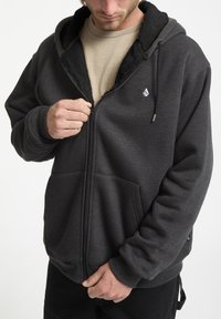 Sort zip-up hoodie med blødt, pelsforet indre, kængurulommer og et lille hvidt logo på brystet. Casual design med ribbede manchetter.