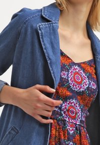 Veste en denim avec un léger délavage, col à revers et poches avant, portée sur une robe à motifs floraux aux couleurs vives.