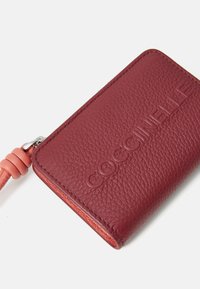 Portafoglio rosso in pelle con finitura testurizzata, con logo in rilievo e una fascia in pelle rosa con un nodo decorativo, chiusura a zip.