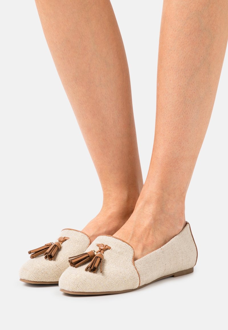 Mocassins tissés beige avec des pampilles marron portés aux pieds sur un fond clair uni.
