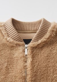 Giacca bomber in finta pelliccia color beige con colletto a coste. Chiusura frontale con zip e texture morbida, ideale per un look casual nelle giornate fresche.