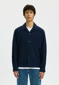 Chemise boutonnée bleu marine avec une poche, fabriquée en tissu léger. Elle présente un col à pointes écartées et des manches longues, associée à un jean bleu.