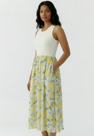 Frau mit langen welligen schwarzen Haaren, die ein ärmelloses weißes Oberteil und einen hellblauen Rock mit gelbem Zitronen- und grünem Blätterdruck trägt, Hände in den Taschen.