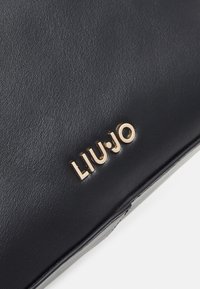 Surface en cuir noir avec des lettres métalliques dorées formant le mot "LIU•JO" fixées près du bord inférieur.