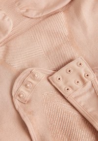 Nærbillede af beige stof med teksturerede mønstre og trykknapper på elastiske stropper for justerbar pasform.