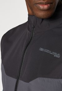 Schwarz- und graue Radjacke mit hohem Kragen, Reißverschluss und dezentem Branding. Der Stoff wirkt glatt mit matter Oberfläche.