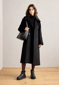 Abrigo largo de lana negra con amplias solapas y parte delantera de doble botonadura. El modelo lleva un suéter negro de cuello alto, un bolso negro y botines negros con estampado.