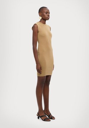 Robe moulante en tissu tricoté beige, avec un col montant et des manches courtes. Associée à des sandales noires à lanières. Texture lisse, design ajusté.
