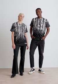 Zwart-witte graphic t-shirts met ingewikkelde patronen; gemodelleerd met zwarte broek en sneakers. Het ontwerp bevat abstracte lijnen en vormen.