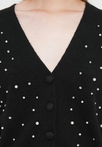 Svart cardigan med V-hals, med en tekstur av stoff og dekorative elementer som perler og rhinestones spredt over forsiden.