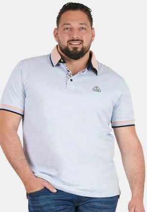 Homme barbu souriant, portant un polo bleu clair avec un col à motif et un jean bleu, main dans la poche, sur fond blanc uni.