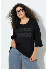 Zwart langermelig T-shirt met een ronde hals en de tekst "LOOK GOOD" in een bladpatroon; gecombineerd met lichtblauwe spijkerbroek. Zacht, gladde stof.