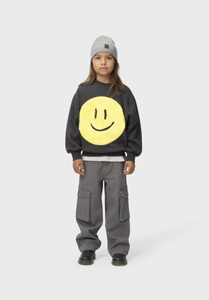 Enfant portant un bonnet gris, un sweat-shirt noir avec un grand smiley jaune, un pantalon cargo gris et des bottes noires, debout sur un fond uni.
