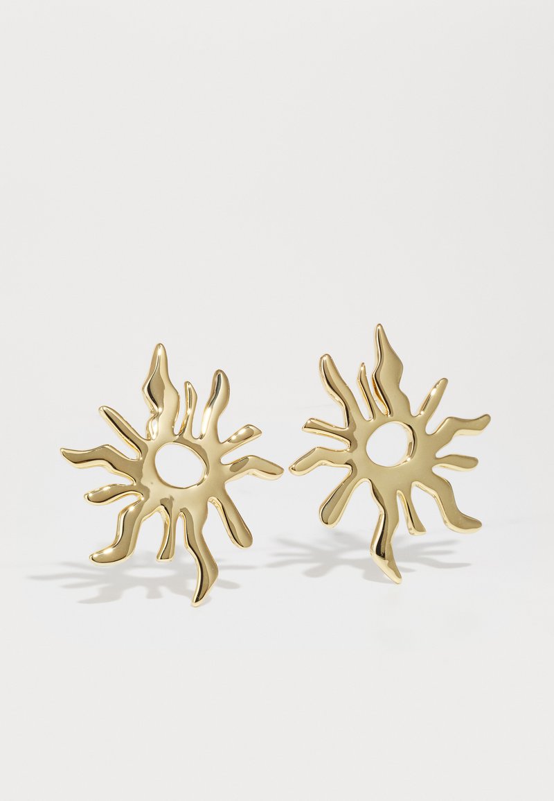 Gold earrings shaped like suns, featuring a textured surface, pointed rays, and circular cutouts in the center. Shine and detail are prominent.
