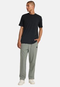 Black short-sleeve t-shirt, relaxed fit, paired with light green sweatpants featuring a side logo patch. Both garments made of soft fabric.