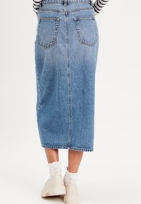 Jupe midi en denim de couleur bleu clair avec une coupe droite. Elle possède deux poches arrière et des passants de ceinture. Portée avec des chaussettes blanches et des chaussures beiges imposantes.