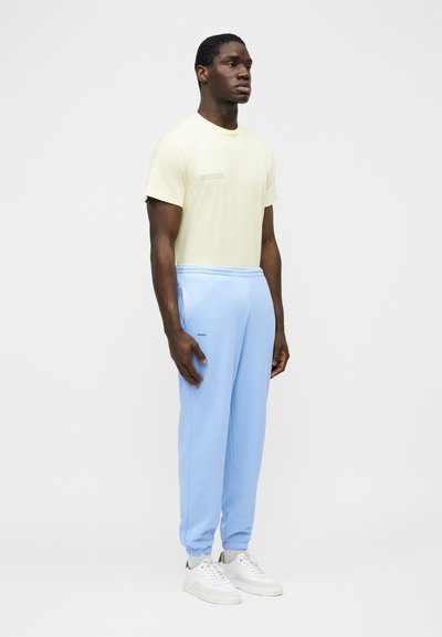 Fekete férfi áll jobbra nézve, világos sárga rövid ujjú ingben, világoskék sportnadrágban és fehér tornacipőben, egyszerű világosszürke háttér előtt.