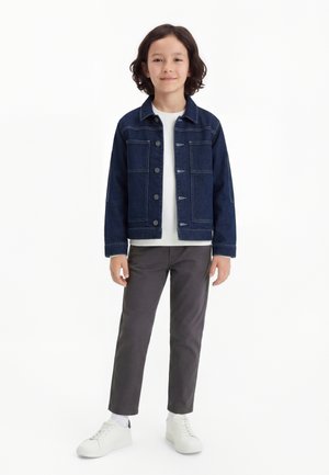 Dziecko o włosach do ramion, ubrane w ciemnoniebieską kurtkę dżinsową, białą koszulę, szare spodnie i białe trampki, stojące na białym tle.