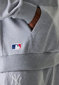 Grå sweatshirt med ribbede manchetter, stor frontlomme og MLB-logo patch. Stoffet har en glat tekstur og en afslappet pasform.