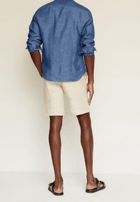 Blaues Chambray-Hemd mit hochgekrempelten Ärmeln, beiger Shorts und braunen Flip-Flops, die ein entspanntes Sommeroutfit von hinten zeigen.