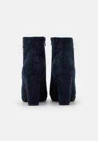 Botas de tobillo de ante azul marino con un alto tacón cuadrado, cierre de cremallera y adornos de perlas decorativas en el costado. Textura suave.