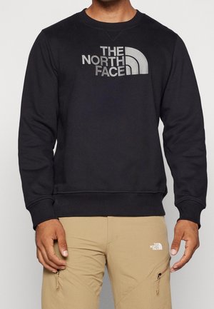 Mand iført sort sweatshirt med stort "The North Face" logo og beige bukser med mindre matchende logo nær lommen.