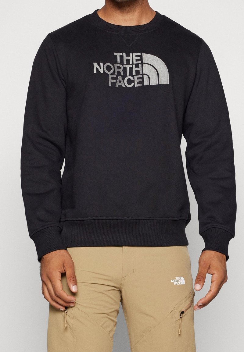 Homme portant un sweat-shirt noir avec un grand logo « The North Face » et un pantalon beige avec un petit logo assorti près de la poche.
