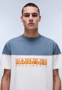 Camiseta de manga corta con sección superior azul y parte inferior blanca. Presenta el logo bordado en naranja "NAPAPURI geographic" en el pecho.