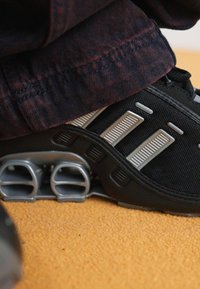 Black sports shoe with gray accents, textured mesh upper, and unique spring-like sole. Visible tread and subtle reflective details.